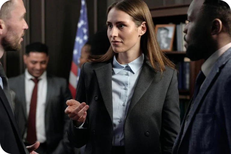 A woman in a dark blazer and light blue shirt speaking to two men in suits in a professional room with an American flag.
