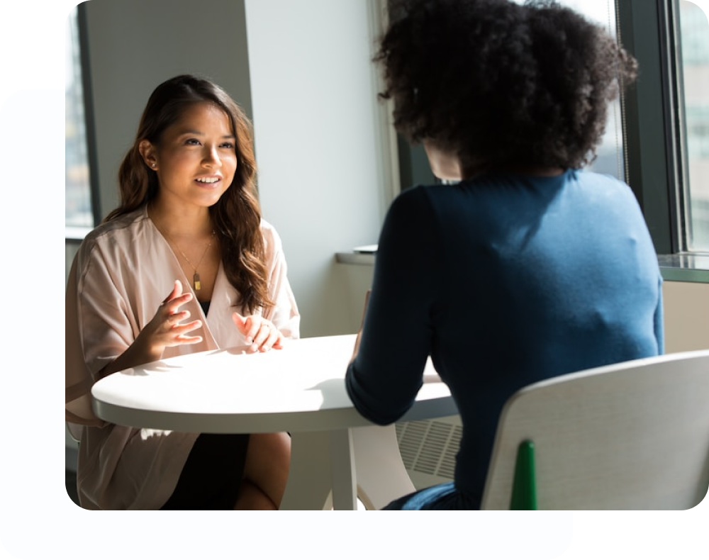 Two women sitting at a white round table, with one woman speaking and gesturing while the other listens from behind.