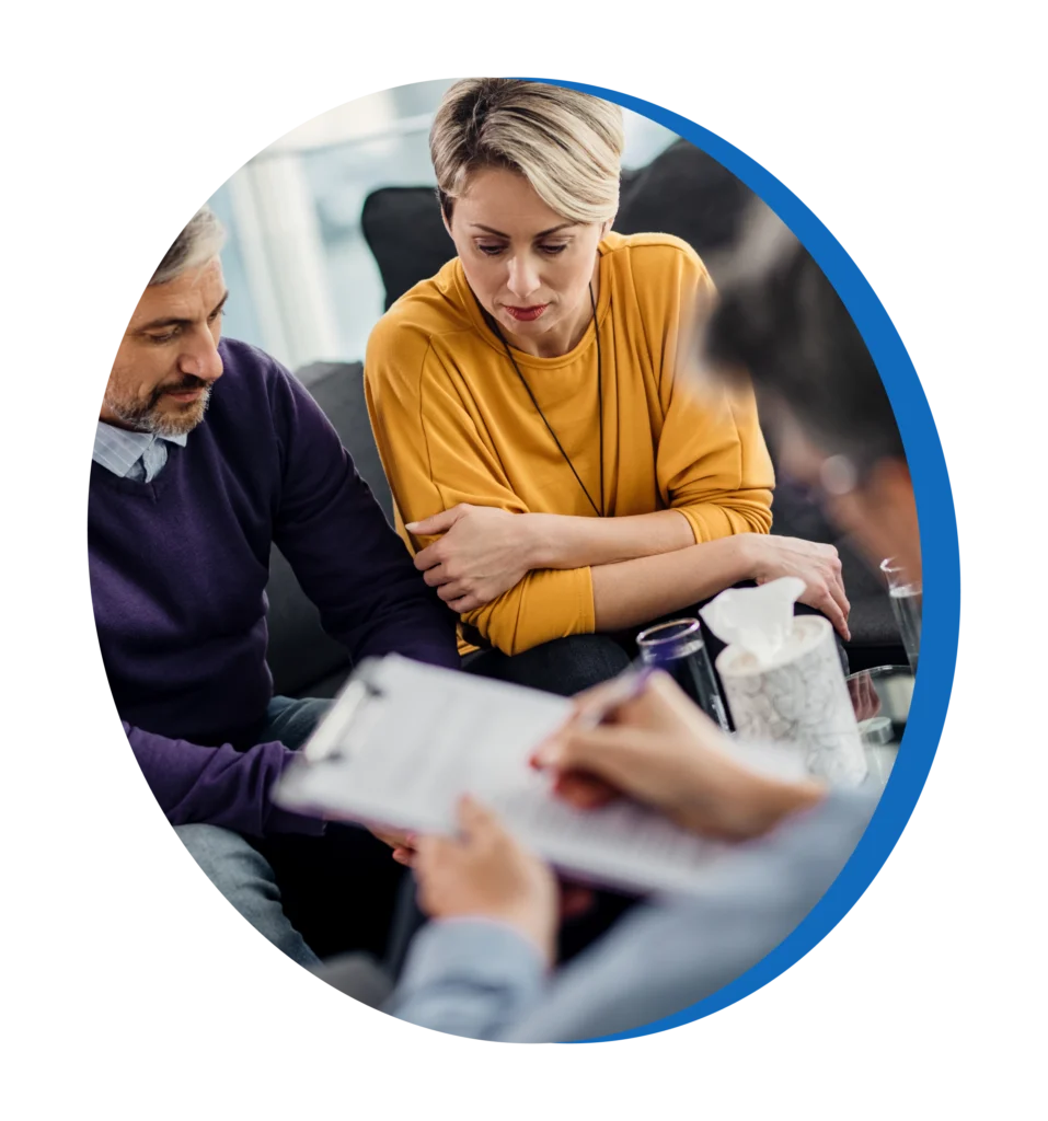 A man and a woman sitting on a sofa, looking down at a clipboard being held by a third person in the foreground.