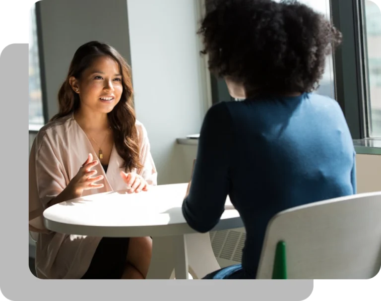 Two women sitting at a white round table, with one woman speaking and gesturing while the other listens from behind.